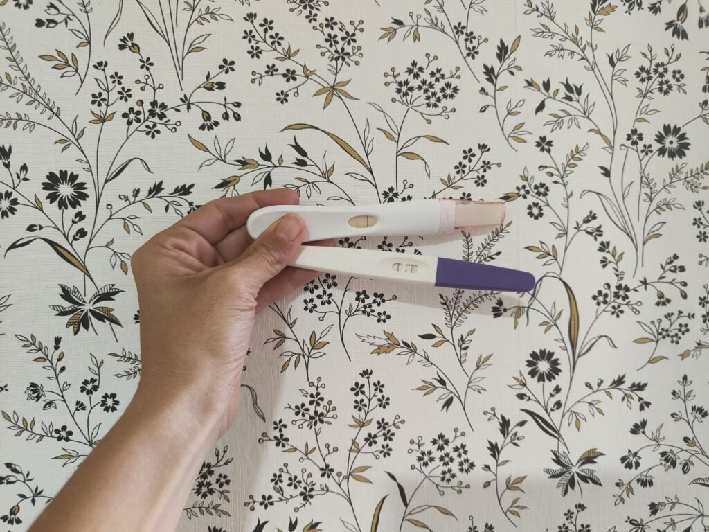 Mano sujetando dos test de embarazo positivos sobre fondo floral, imagen para Mamá Zombi Chic.